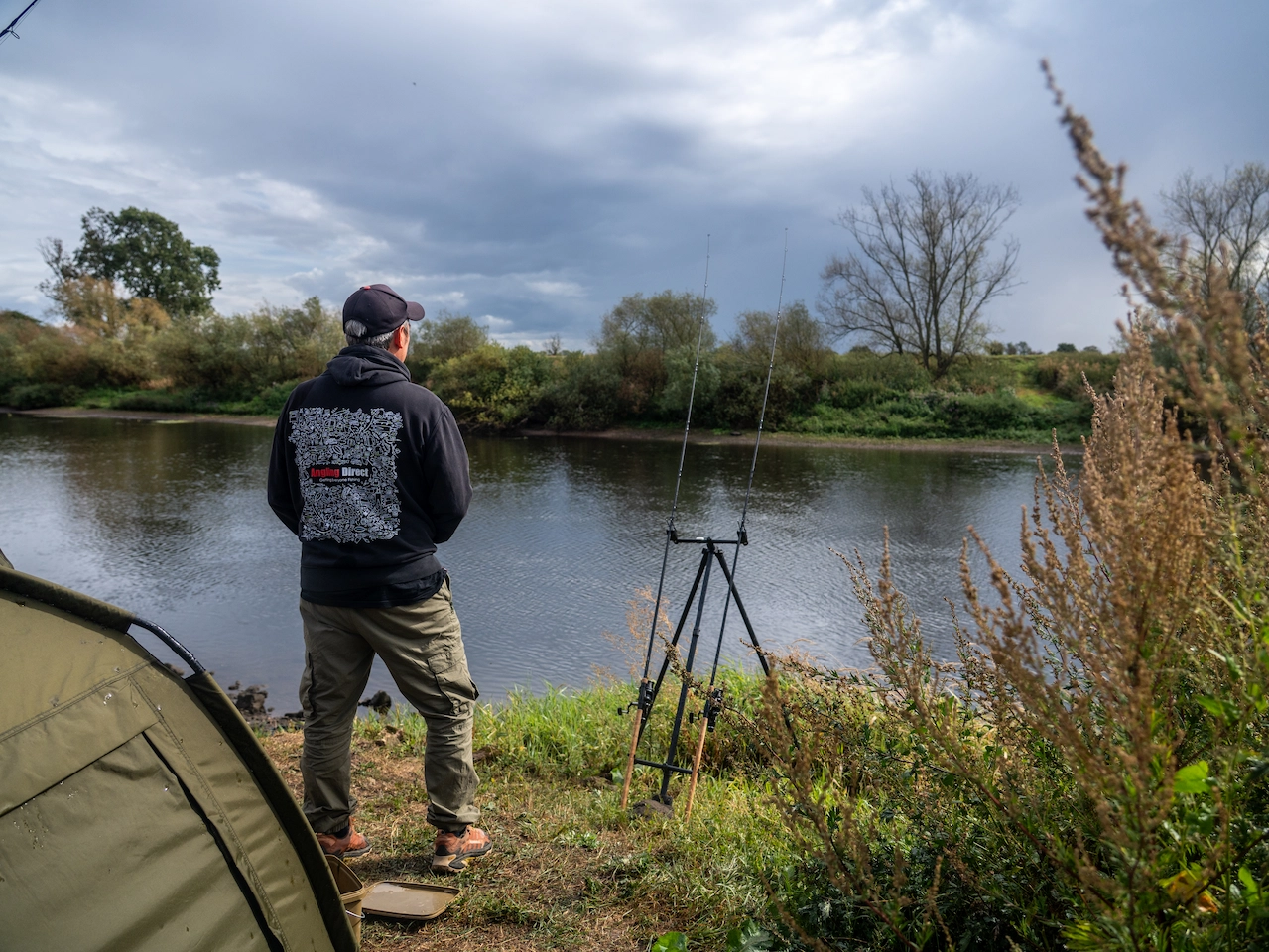 Angler in black Angling Direct hoodie, with two rods setup in front, a bivvy in the foreground. He is looking out across a lake.